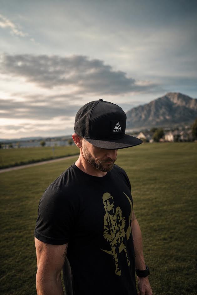 Man wearing The Initiative black flat-brim snapback outdoors at sunset, mountain and open field behind him